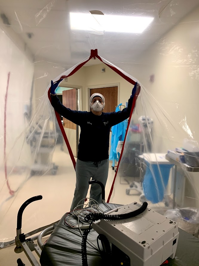 A healthcare worker wearing a mask and gloves stands inside a transparent barrier, holding it open, with medical equipment in the foreground.