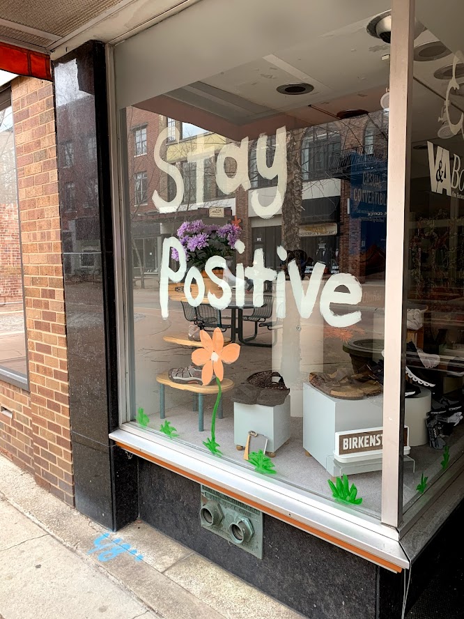 Storefront window with the words 'Stay Positive' painted in white, alongside a drawing of a flower and green grass.