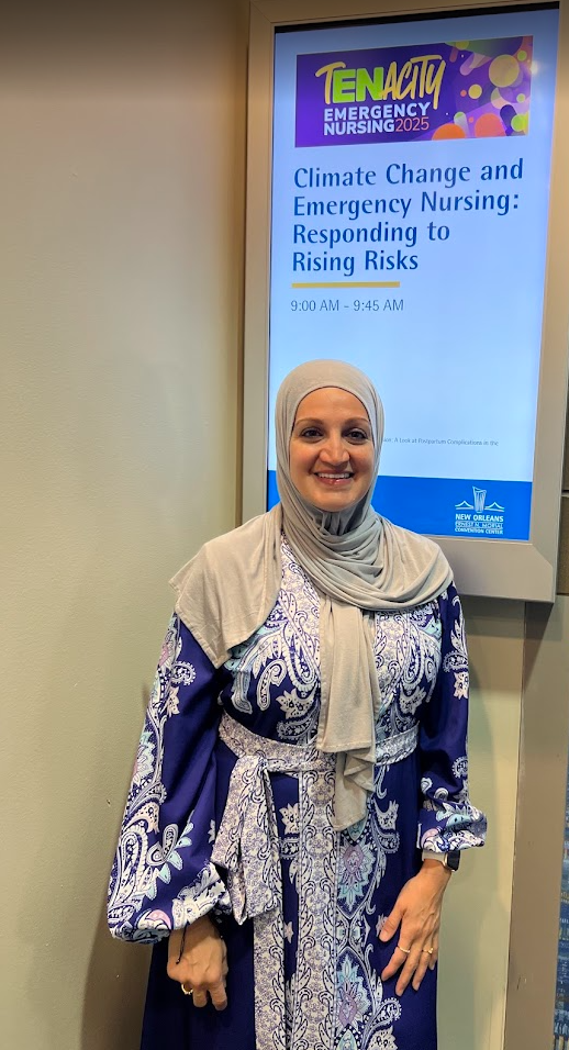 A woman in a blue and white patterned dress with a gray scarf stands in front of a sign for the ENA 2025 conference, featuring the title 'Climate Change and Emergency Nursing: Responding to Rising Risks'.